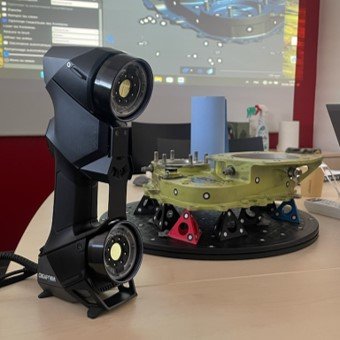 Scanner 3D portable posé sur une table devant une pièce mécanique et un écran